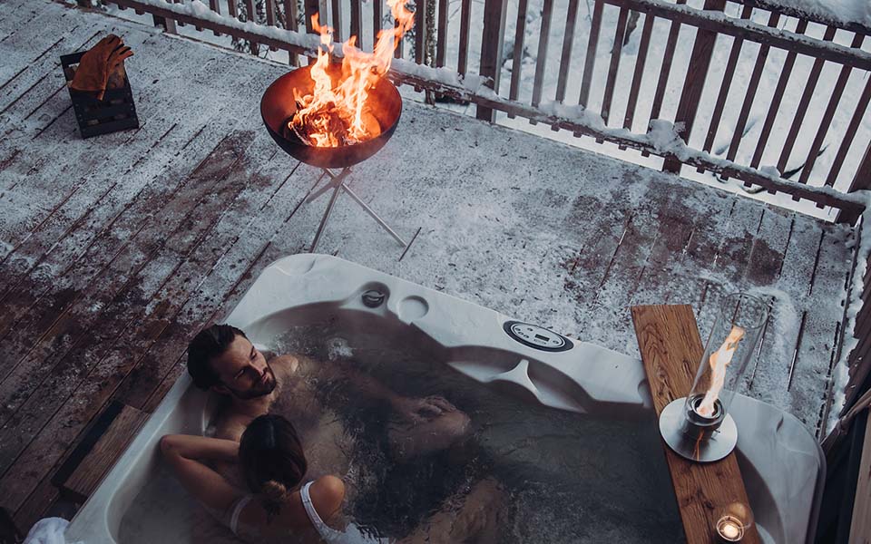An inviting scene with a woman sitting in a hot tub with a fire in the background, highlighted by the presence of the Kurzurlaub Vor Weihnachten Triple2.