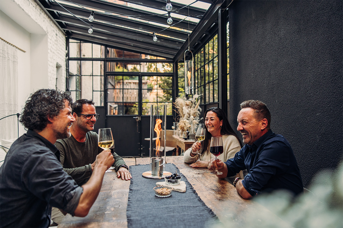 SPIN 1200 Tabletop Fireplace silver Three men sitting at a table with wine glasses, with the 00695 Spin 1200 Tablefire silver adding a functional and elegant touch to the scene.