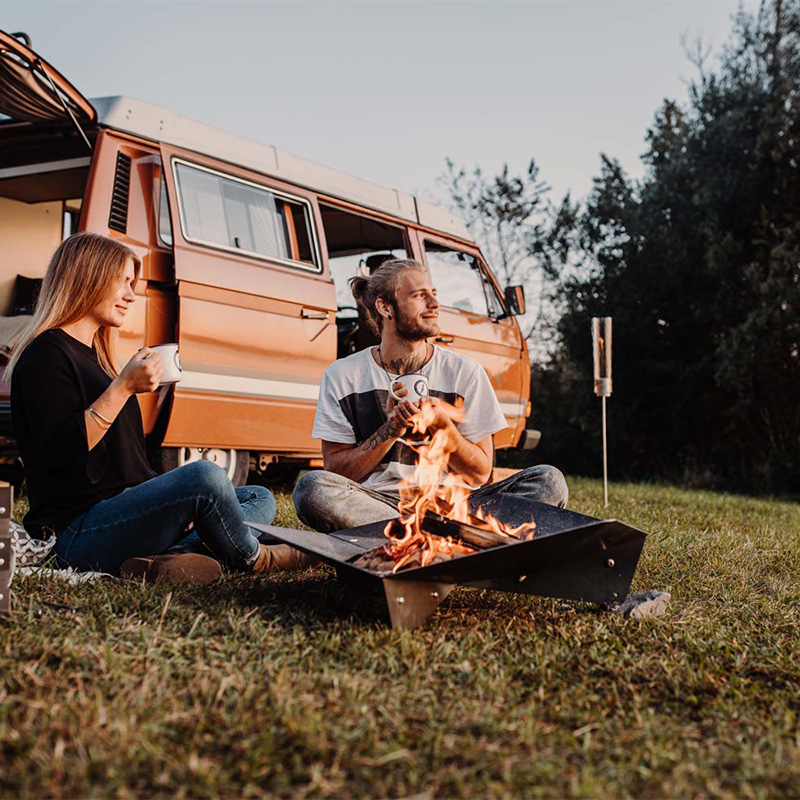 Une scène accueillante avec un couple assis sur l'herbe à côté d'un camping-car, mise en valeur par la présence du TRIPLE Fireplace.