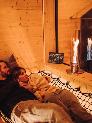 Ein Mann und eine Frau liegen in einer Hängematte vor einem Kamin, bereichert durch die Präsenz des Spin Tischfeuer, das Design mit Funktionalität verbindet.