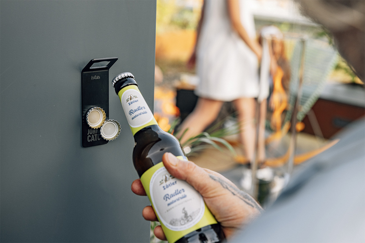 JOHNNY CATCH Magnet Bottle Opener black A person holding a bottle of beer near a door, enriched by the presence of the 00841 Johnny Catch Magnet Black Bottleopener, which merges design with utility.