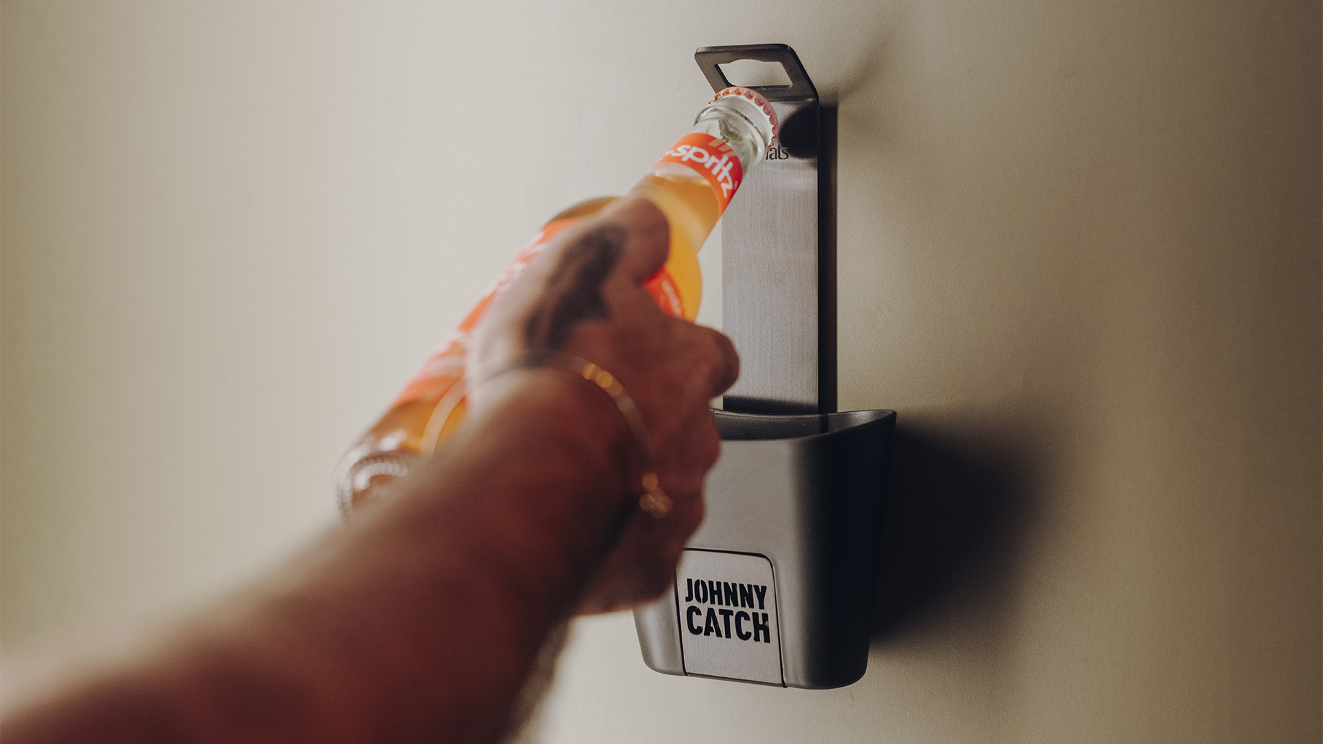 Eine Darstellung einer Hand, die eine Flasche Orangensaft hält, die mit einem Flaschenöffner geöffnet werden soll, wobei der 00842 Johnny Catch Cup Black Flaschenöffner als zentrales und zweckmäßiges Element dient.