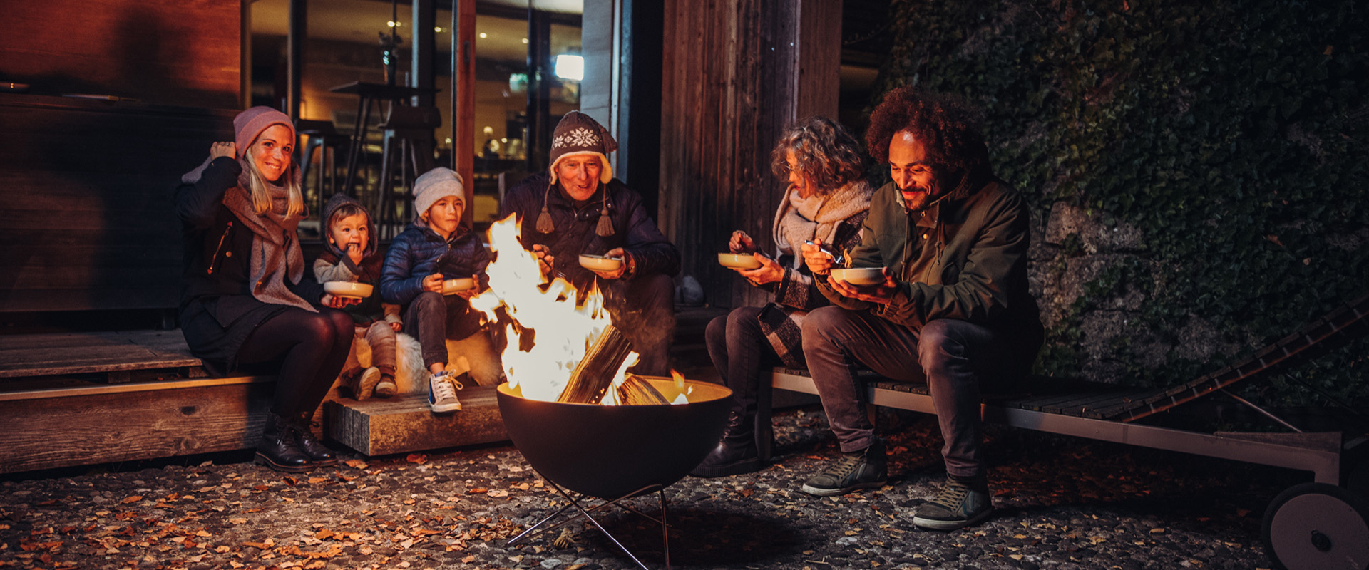 De foto toont een groep mensen die rond een vuur zitten, met de FIRE KITCHEN Reserveplank onderaan zwart.