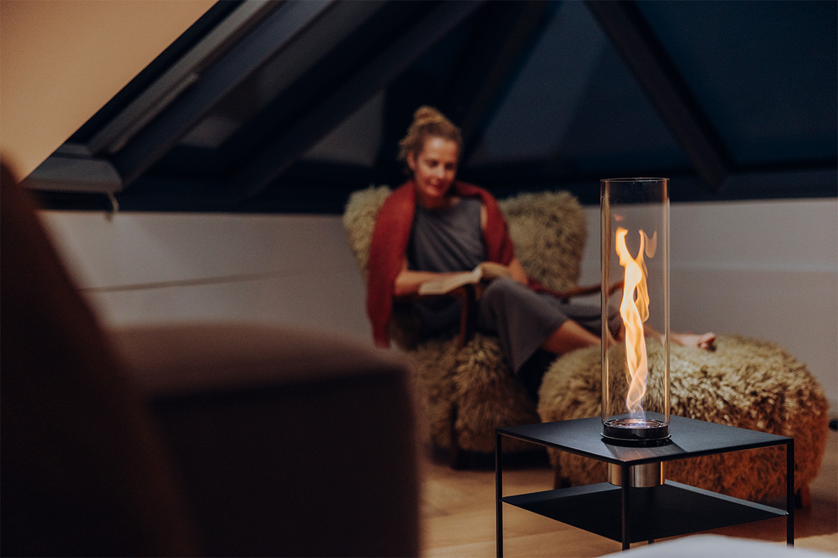 Eine Frau sitzt auf einem flauschigen Sessel und blickt in die Flamme von SPIN air 1200 Einbau, eingebaut in den FIRE FRAME Lounge-Tisch.