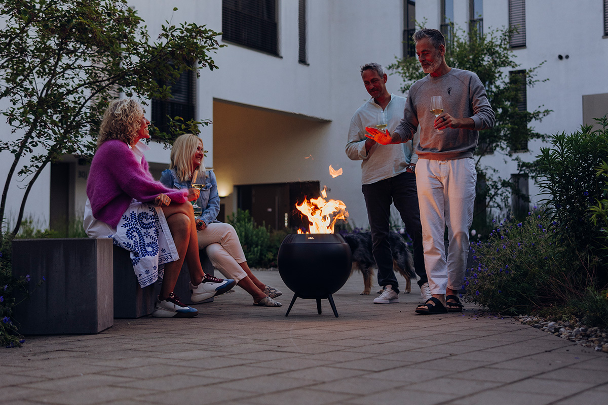 MOON 45 all black Feuerkorb mit niedrigem Fuß Vier Personen sitzen und stehen um die wärmende Feuerschale MOON.