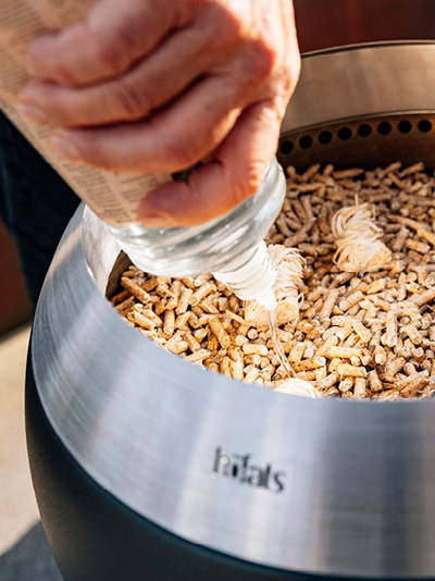 A person is pouring ethanol over wooden chips into a fire basket. Showing how to light the MOON 45
