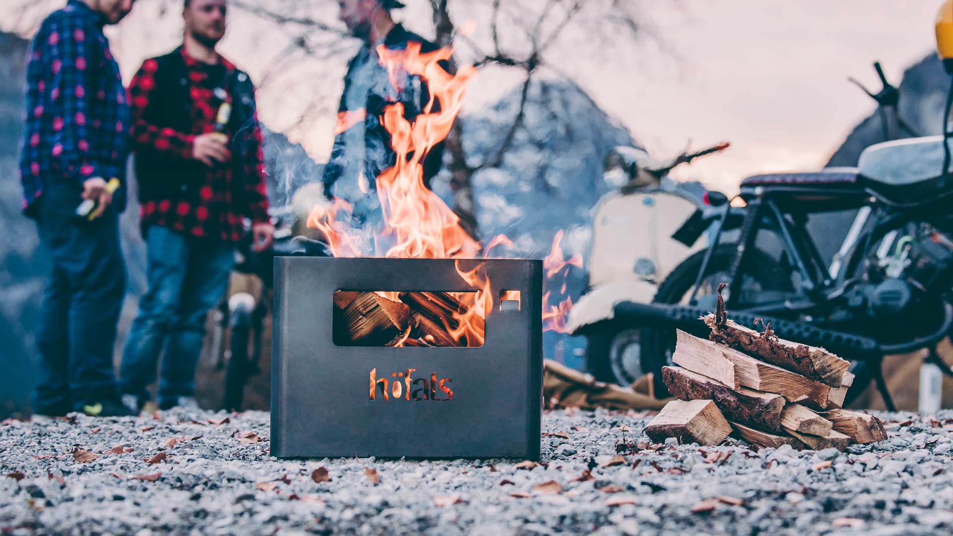 A campfire with people standing around it the Beer Box Firebasket complements the moment with its thoughtful design.
