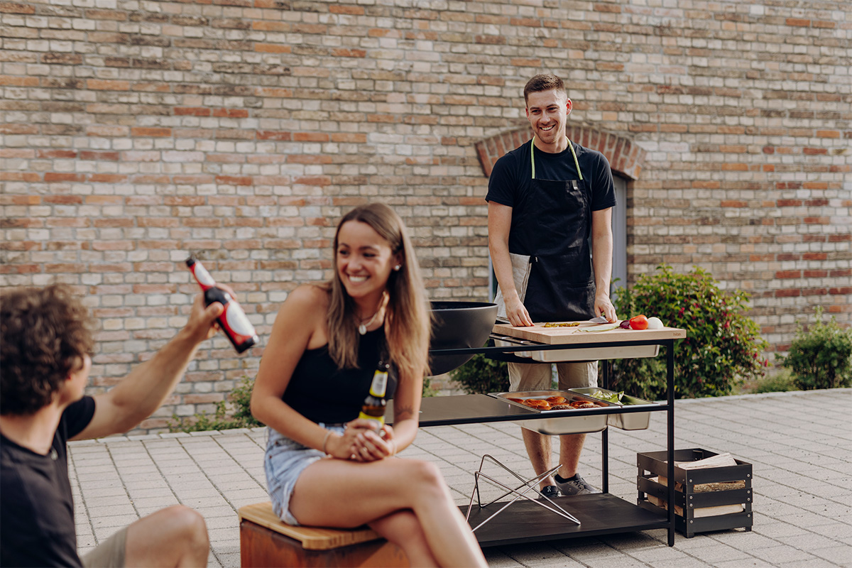 FIRE KITCHEN with BOWL 57 Plancha BBQ Set low The photo presents a man and woman sitting on a bench with a grill, with the FIRE KITCHEN with BOWL 57 Plancha BBQ Set incl. wheels.