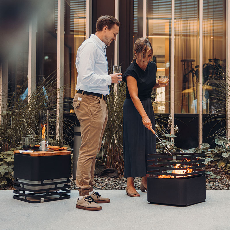 Experience a man and woman standing next to a fire pit with the refined touch of the Cube Firebasket