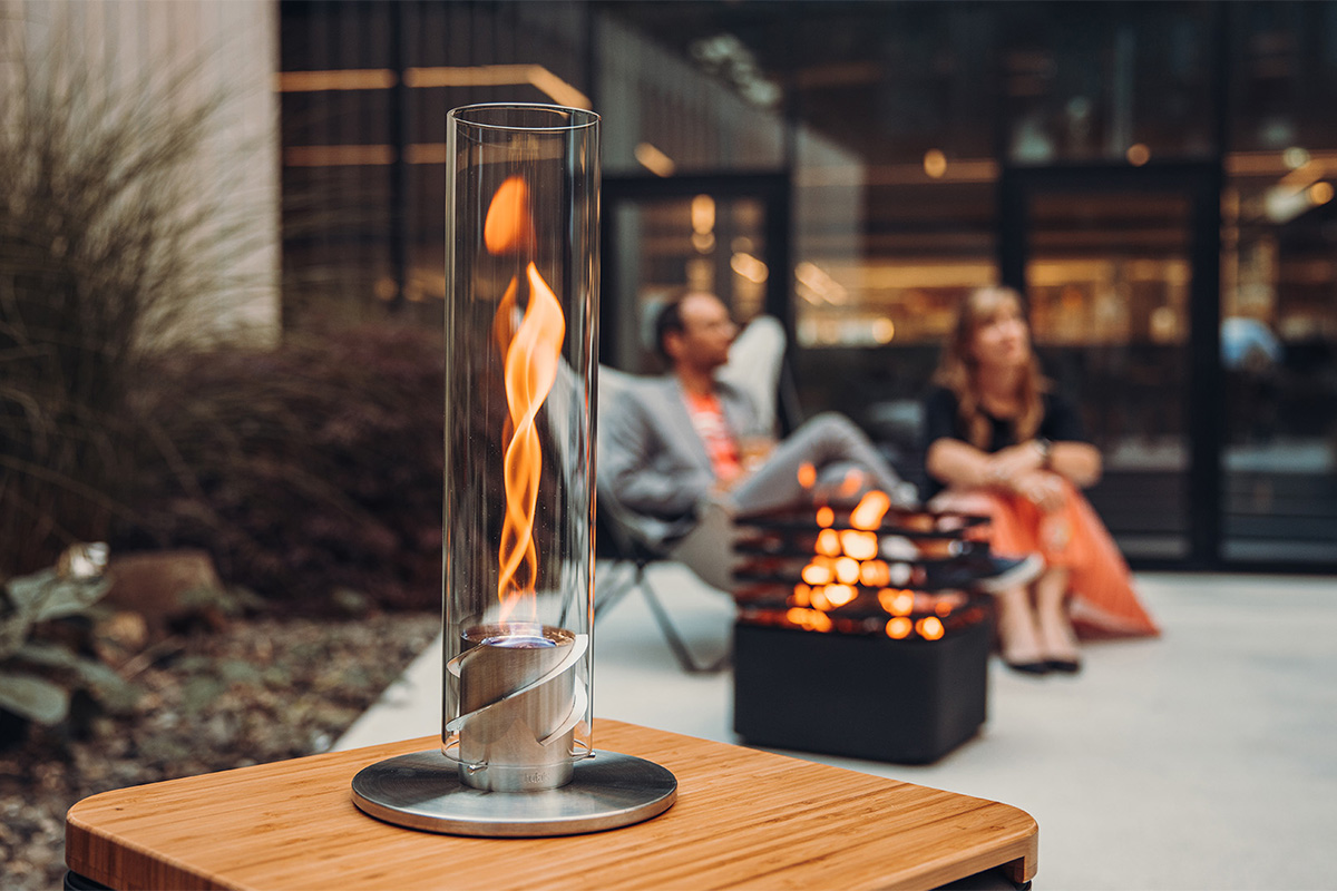 An image showing a fire pit with a couple sitting in the background, with the SPIN 900 Tabletop Fireplace silver.
