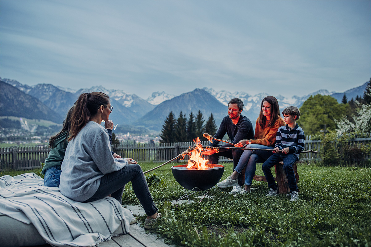BOWL 57 Feuerschale mit Sternfuß Eine Frau und zwei Kinder sitzen um eine Feuerstelle herum, wobei die Schale 00249 Bowl 57 mit Sternständer eine funktionale und elegante Note zur Szene hinzufügt.