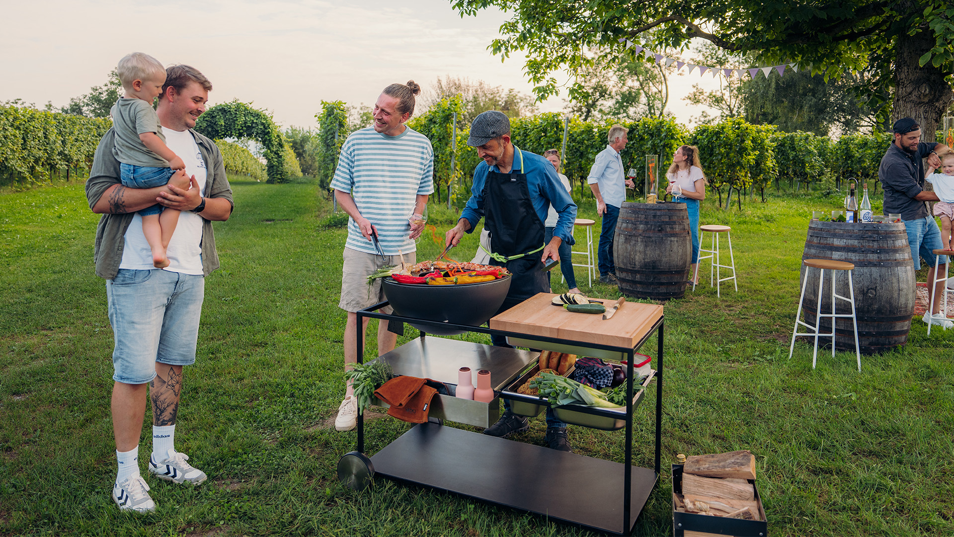A depiction of a group of people standing around a table, featuring the 00739 Fire Kitchen Bowl 57 Plancha Grill Set as a central and purposeful element.