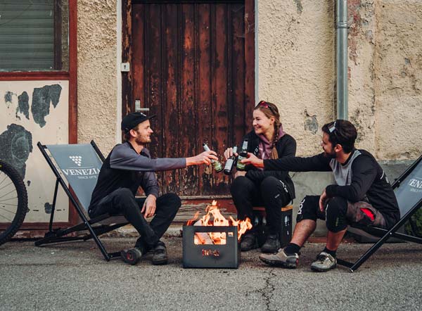 Fire under the saddle The photo presents three people sitting around a fire pit, with the BEER BOX Fire Basket.