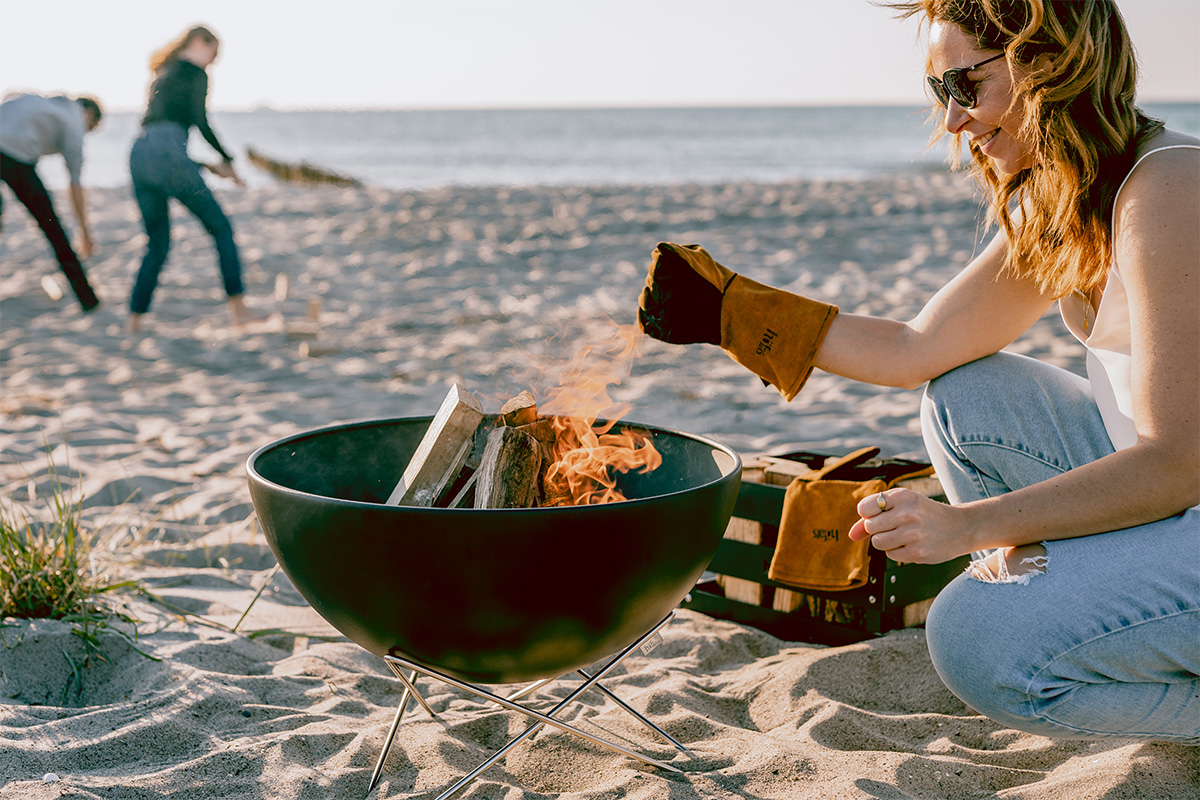 BOWL 57 Feuerschale mit Sternfuß Eine Darstellung einer Frau, die am Strand sitzt und die 00249 Bowl 57 mit Sternständer als zentrales und zweckmäßiges Element zeigt.