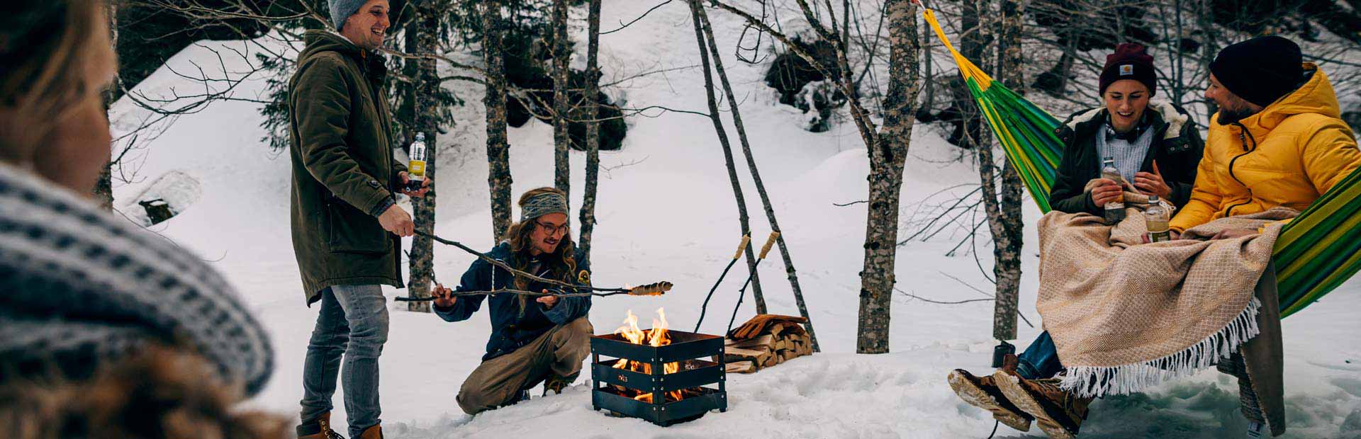 La photo montre des personnes assises autour d'un feu de camp dans les bois, avec le CRATE Fire Basket.