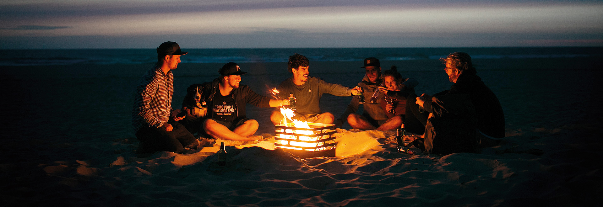 A group of people sitting around a fire on the beach. Showing the Cube firebasket as a central element of the scene