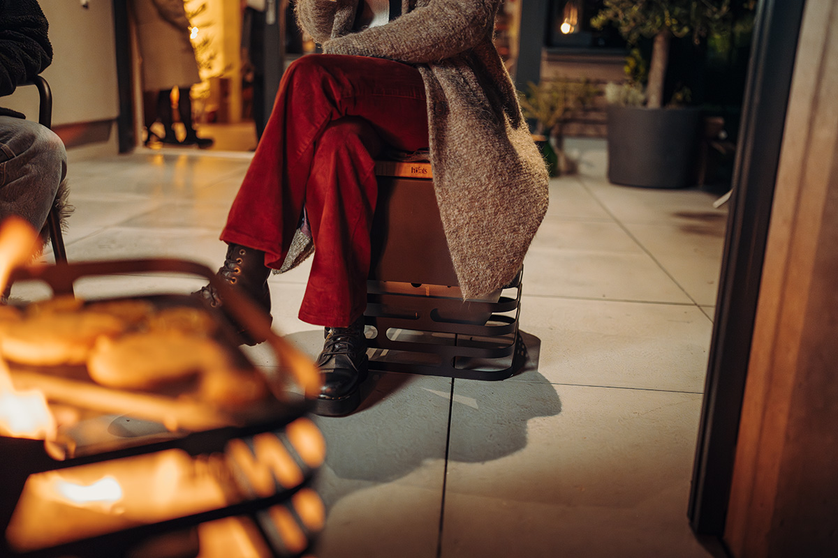 CUBE Flip Set schwarz Eine Darstellung einer Frau, die auf einem schwarzen Kubus-Feuerkorb sitzt, wobei der Cube Flip als zentrales und zweckmäßiges Element dient.