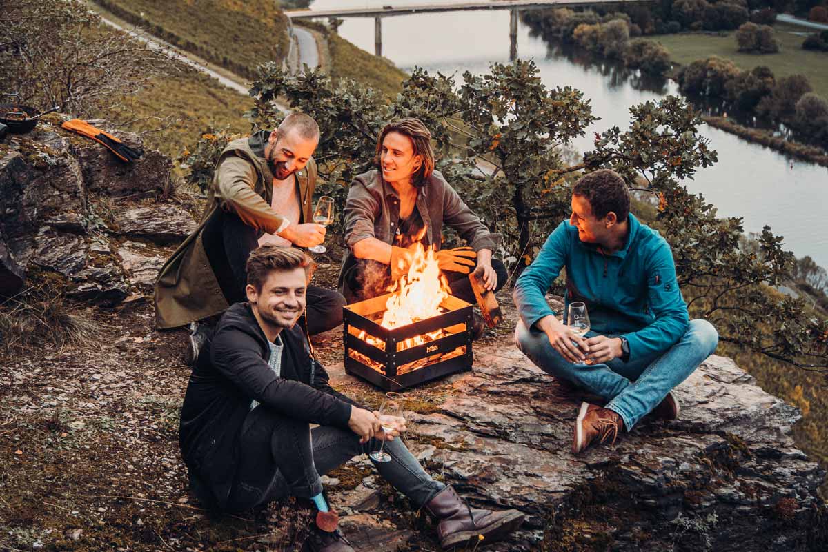 CRATE Feuerkorb Eine einladende Szene mit drei Personen, die um ein Lagerfeuer sitzen, hervorgehoben durch die Anwesenheit des Crate Firebasket.