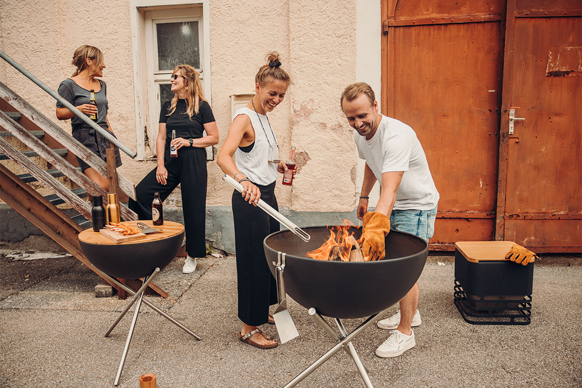 BOWL 70 Bol à Feu avec Trépied Un groupe de personnes se tenant autour d'un brasero, avec le BOWL 70 avec trépied 00258 ajoutant une touche fonctionnelle et élégante à la scène.