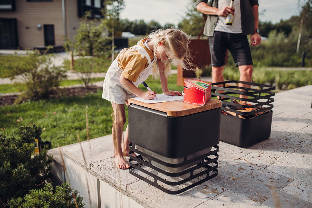 CUBE Flip Set schwarz Ein kleines Mädchen steht auf einem Betonblock und benutzt einen Cube-Feuerkorb als Malunterlage, bereichert durch die Anwesenheit des Cube Flip, der Design und Nutzen vereint.