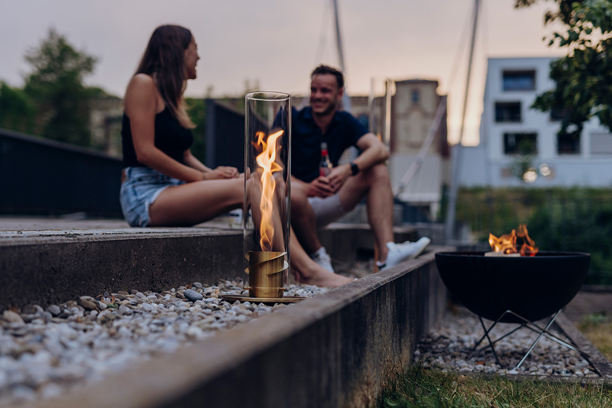 An inviting scene with a man and woman sitting on a bench next to a fire, highlighted by the presence of the 00699 Spin 1200 Tablefire Gold.