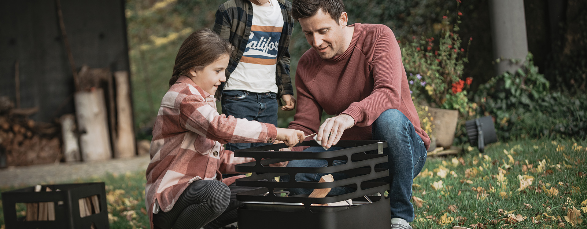 A man and a child are sitting behind a Cube Firebasket, lighting up the fire