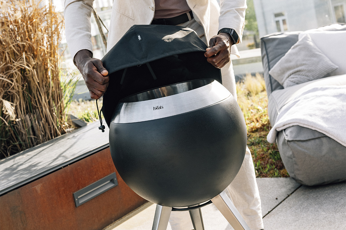 MOON 45 Abdeckhaube Ein Mann hält eine große schwarze Abdeckung über einem Feuerkorb, wobei die 00815 Moon 45 Abdeckung eine funktionale und elegante Note zur Szene hinzufügt.