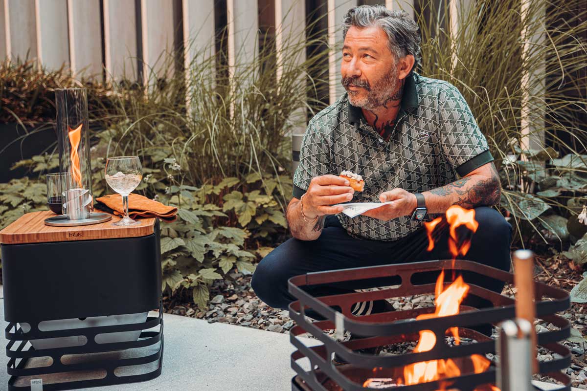 CUBE Flip Set schwarz Eine einladende Szene mit einem Mann, der neben einem Feuerplatz auf dem Boden sitzt, hervorgehoben durch die Anwesenheit des Cube Flip Sets.