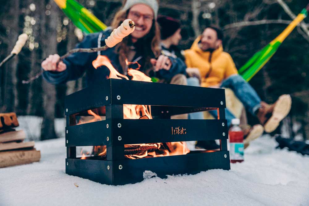 CRATE Feuerkorb Erleben Sie einen Mann und eine Frau, die um ein Lagerfeuer sitzen, mit dem eleganten Touch des Crate Firebasket.