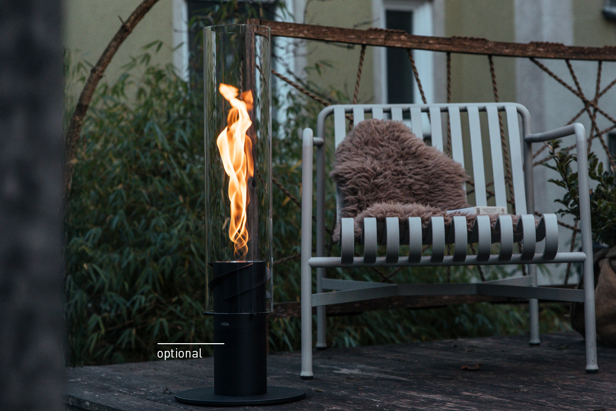 SPIN 1500 Tischfeuer schwarz Ein Feuer brennt in einer SPIN Tischfeuer, draußen neben einem gemütlichen Stuhl, wobei die 00628 Spin 1500 Tischfeuer schwarz der Szene eine funktionale und elegante Note verleiht.
