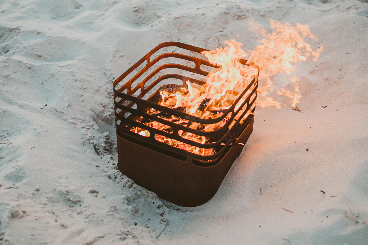 CUBE Feuerkorb rostig Ein Feuerplatz am Strand mit Sand, wobei der Cube Firebasket rostig eine funktionale und elegante Note zur Szene hinzufügt.