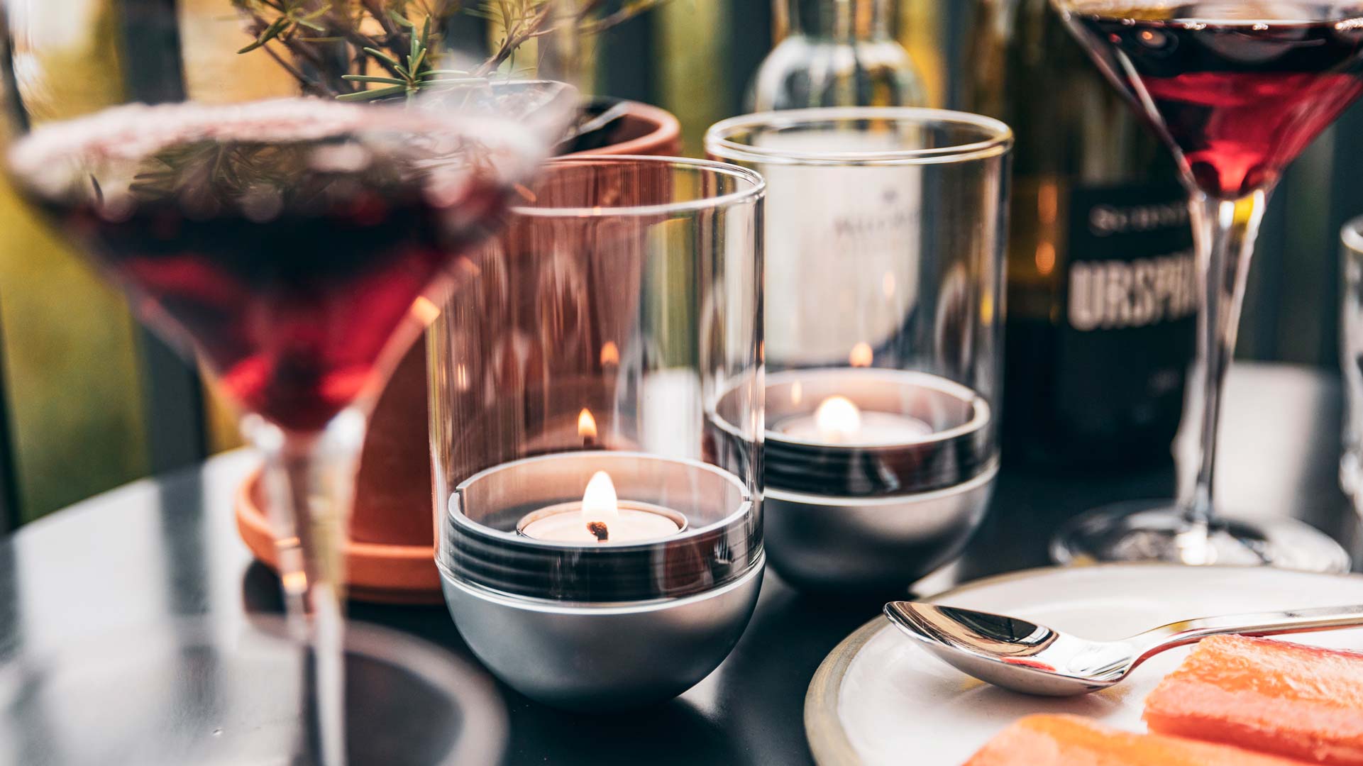 De foto toont een tafel met een kaars, wijnglazen en een bord eten, met de LOGO GRAVITY CANDLE M90 lantaarn.