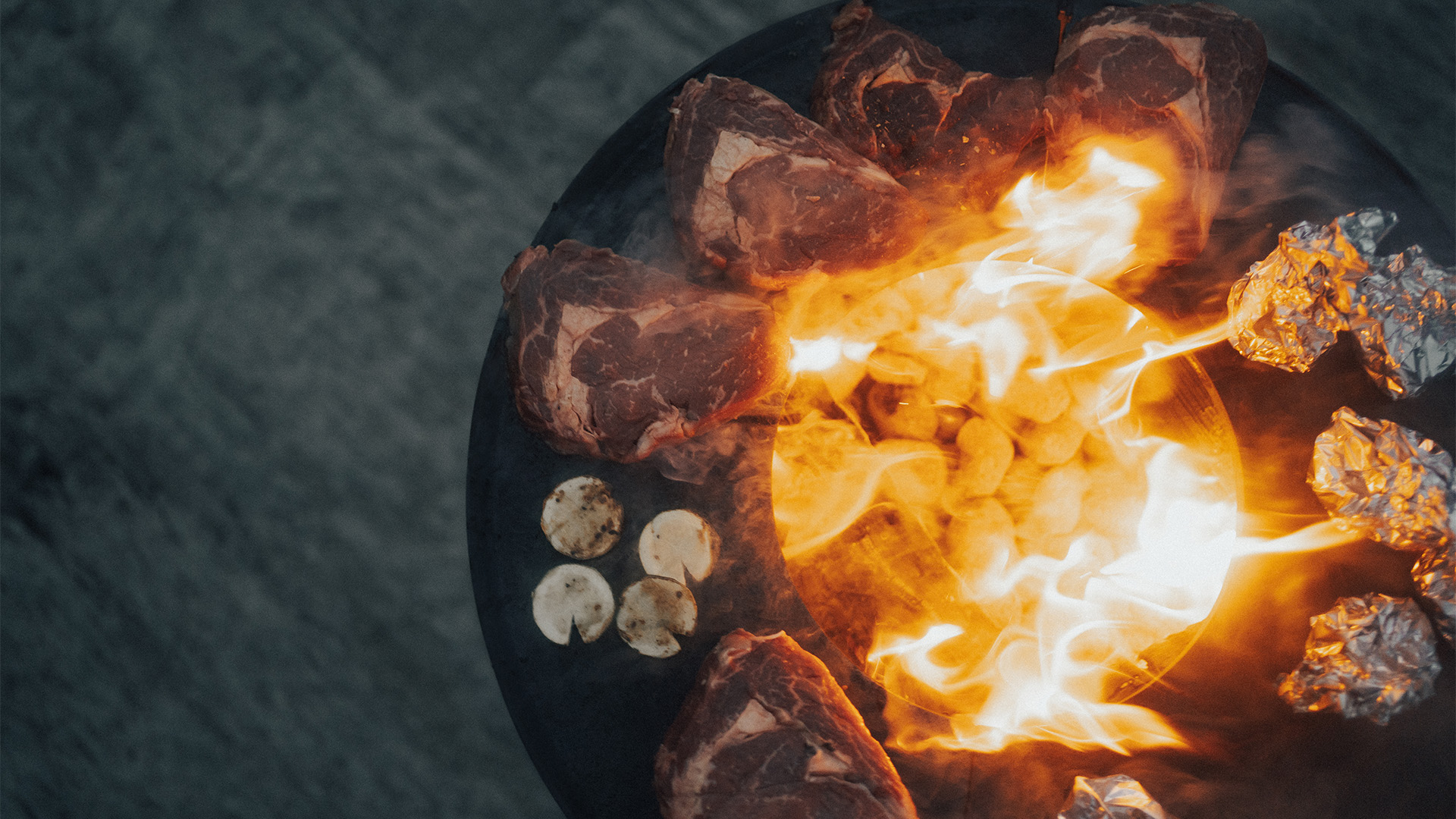 Hier is een vuurkorf afgebeeld met een hoop vlees erop, met de BOWL 57 Plancha BBQ Set laag.