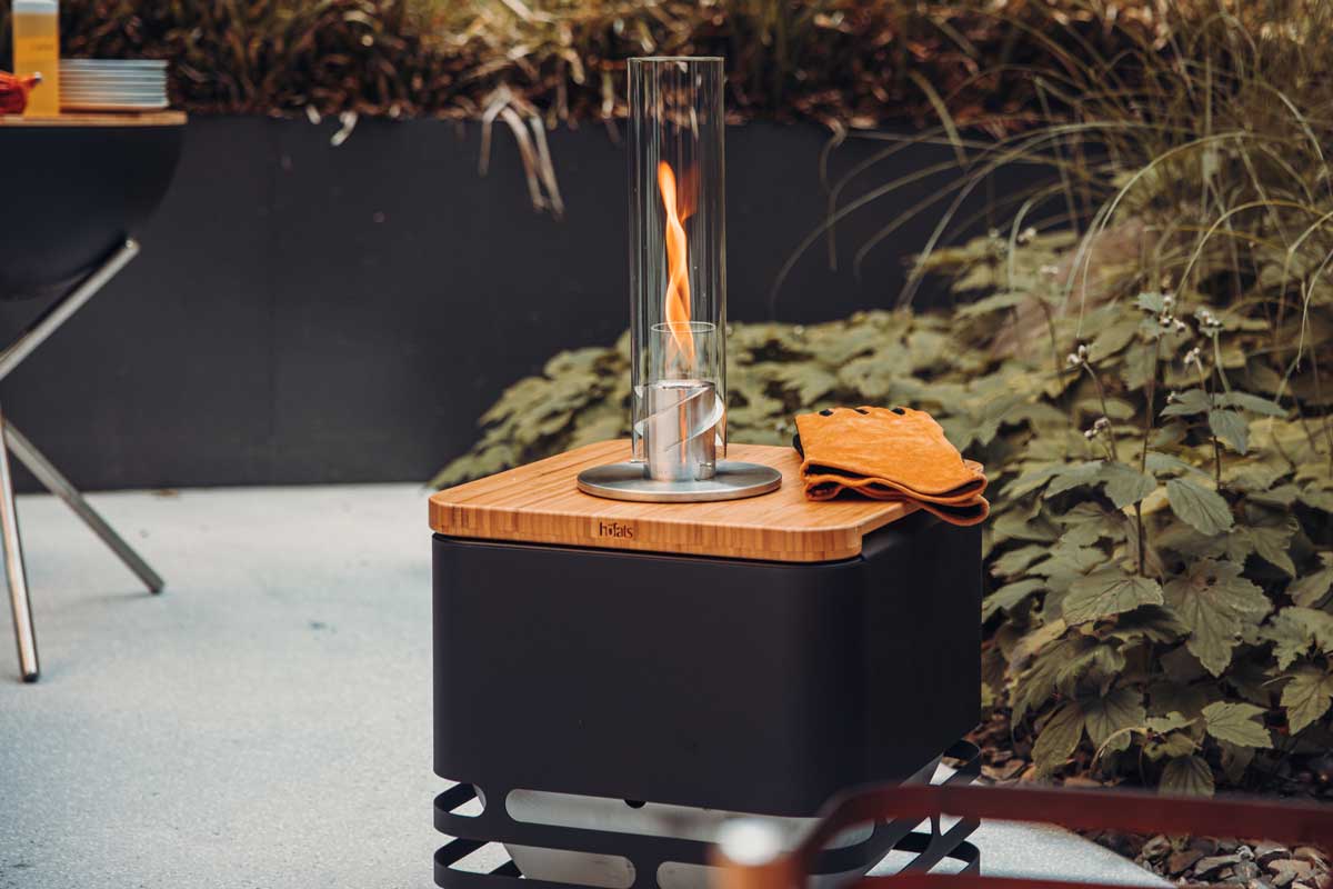 CUBE Flip Set schwarz Ein schwarzer Metallgrill mit einem Holzbrett darauf, auf dem der SPIN Tischfeuer und Lederhandschuhe liegen. Zeigt die Verwendung des Cube Flip Sets.