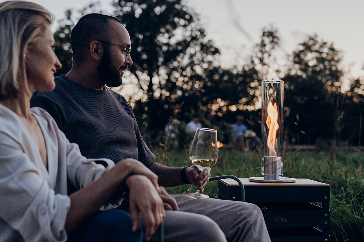 Depicted here is a man and woman sitting on a bench, with the SPIN 900 Tabletop Fireplace silver.