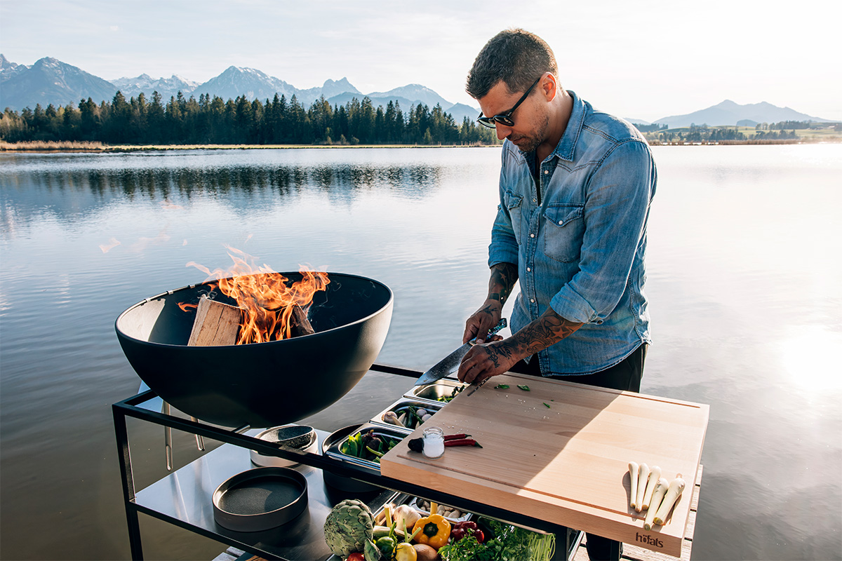 Een man kookt op een grill, waarbij het 00614 Fire Kitchen Worktop een functionele en elegante touch aan de scène toevoegt.