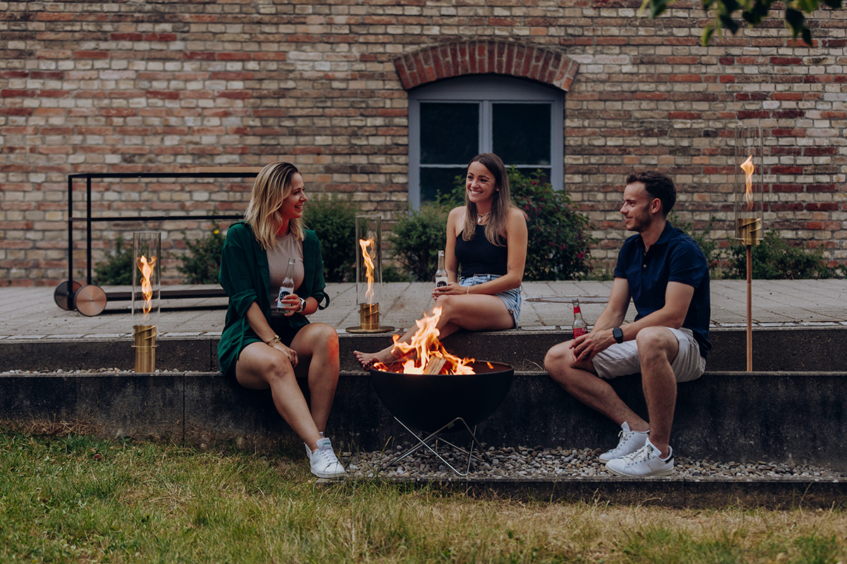 The photo presents a man and a woman sitting on a bench with a fire, with the SPIN 1200 Tabletop Fireplace gold.
