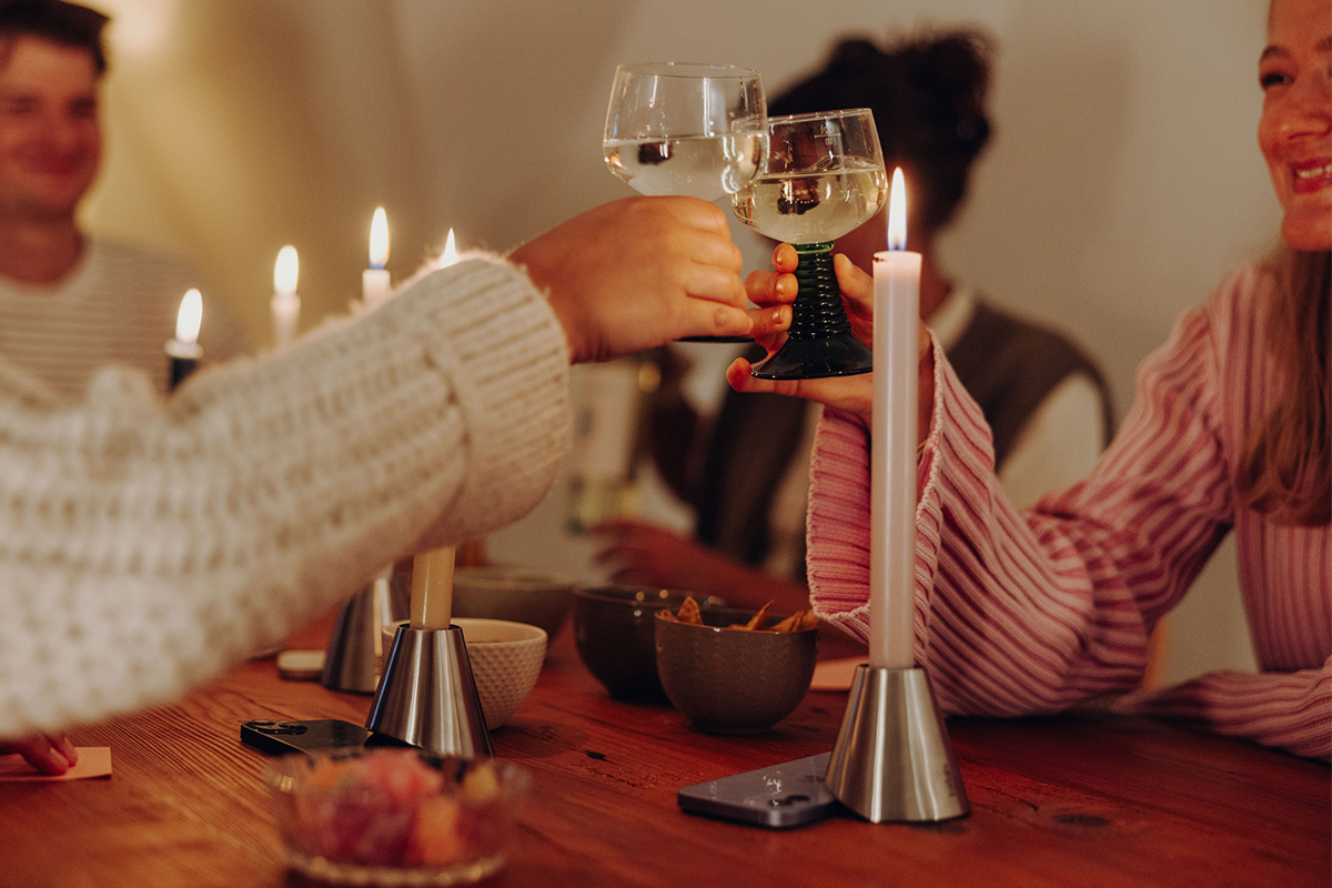 Drei Digi Detox mit Smartphone darunter auf Küchentisch. Daneben zwei Hände die mit einem Weinglas anstoßen.