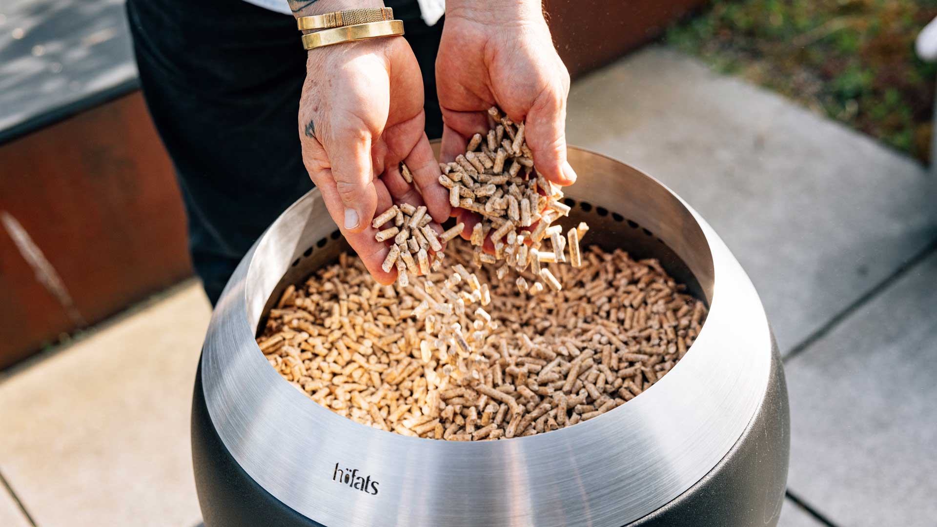Ein Bild zeigt eine Person, die eine Handvoll Holzstücke in einen Metallbehälter wirft, wobei die 01003 Premium Holzpellets als zentrales und zweckmäßiges Element dargestellt sind.