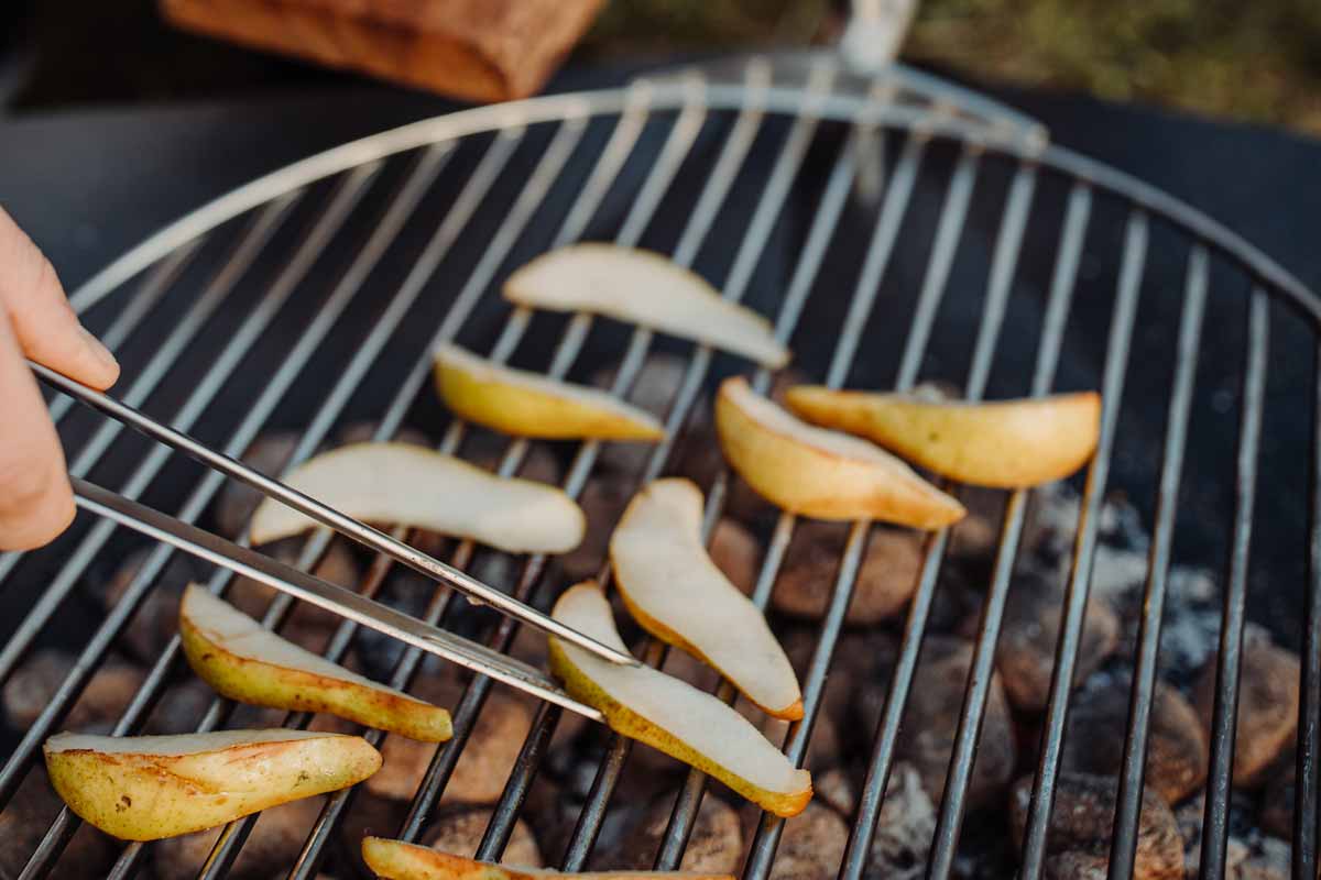 Iemand gebruikt een tang om de aardappelen van de grill te halen, terwijl de Triple 65 openhaard BBQ-set een functionele en elegante touch aan de scène toevoegt.
