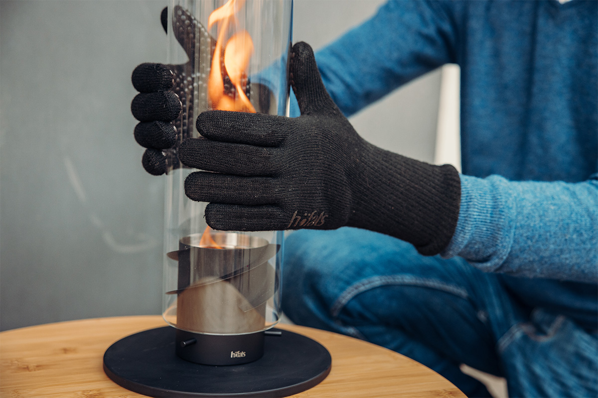 Feuerhandschuhe Aramid Das Bild zeigt eine Person in einem blauen Pullover und schwarzen Handschuhen, die einen Tischkamin mit Feuer in den Fire Gloves Aramid hält.