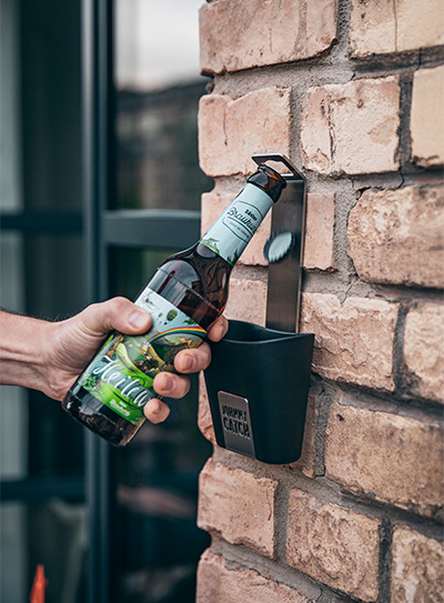 Das Bild zeigt einen Mann, der mit dem JOHNNY CATCH Cup eine Flasche Bier von einer Wand öffnet.