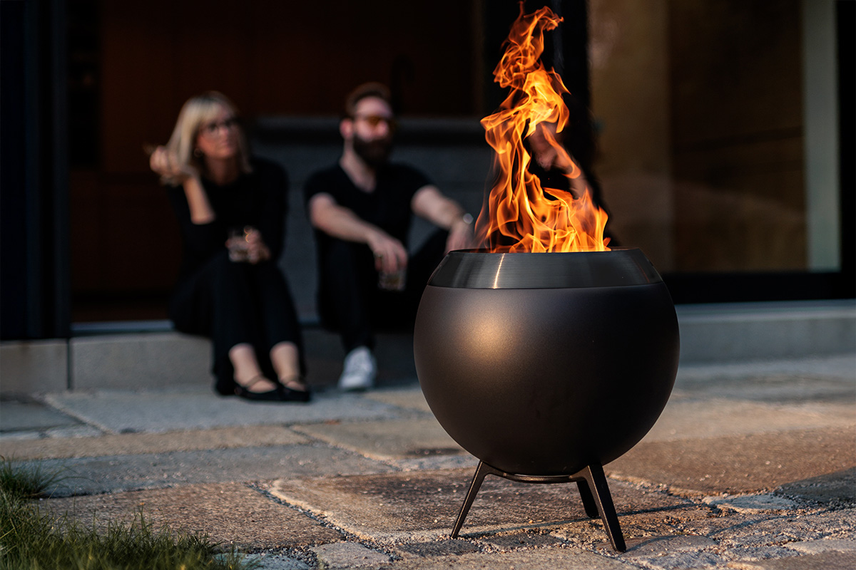 MOON 45 all black Feuerkorb mit niedrigem Fuß Im Vordergrund ist MOON mit niedrigem Fuß zu sehen im Hintergrund zwei schwarz gekleidete Personen, die ins Feuer schauen.