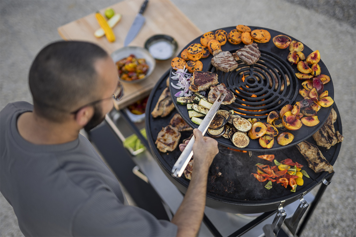 BOWL 70 Warmhaltering Das 00617 Bowl 70 Wärmerelement wird gezeigt, während ein Mann auf einem großen Grill mit einer Zange grillt, was ein stilvolles und zugängliches Bild ergibt.