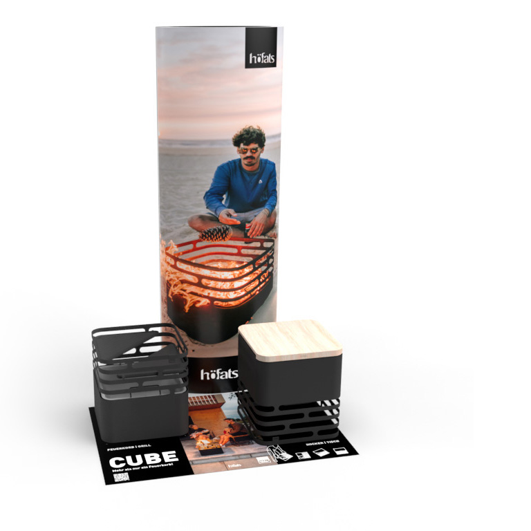 Een kartonnen zuil met een man op het strand naast een vuurplaats en een tapijt met twee Cube-vuurkorven. Toont de Cube-POS-set.