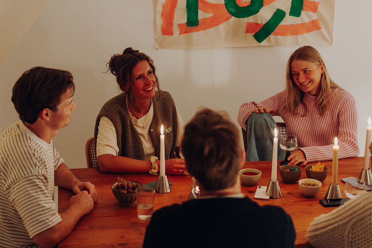 DIGI DETOX Kandelaar (Set van 2) Fünf Personen sitzen ins Gespräch vertieft an Esstisch. Dabei liegen die Smartphones jeweils unter einem Digi Detox Kerzenhalter.